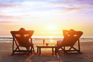 Coucher de soleil aux couleurs rose et doré sur une plage des Landes, avec deux transats installés sur le sable. Deux personnes assises confortablement, profitant d'un moment de détente en mode chill face à l'océan, sous un ciel aux teintes douces et lumineuses, avec des dunes et une végétation naturelle en arrière-plan.