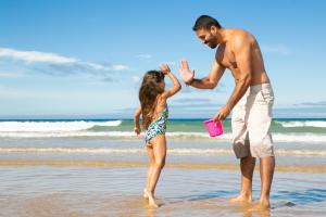 Un père en short fait un high five avec sa jeune fille en maillot de bain, les pieds dans l'eau à la plage. Il tient dans une main un seau rose de château de sable, ils partagent un moment de joie et complicité au bord de l'eau.
