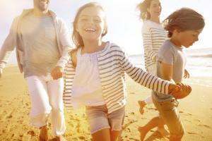 Famille composée d’un papa, d’une maman et de deux enfants courant joyeusement sur le front de mer au coucher du soleil, avec l’océan calme en arrière-plan et un ciel aux teintes orangées, illustrant un moment de bonheur et de complicité en plein air lors d’une soirée d’été.