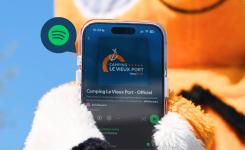 Mascotte du camping représentant un personnage souriant et convivial, tenant un smartphone ouvert affichant la playlist Spotify du camping avec plusieurs chansons en cours de lecture, illustrant l'ambiance musicale et la convivialité du lieu