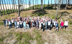 Vue aérienne réalisée par drone d’un groupe d’environ 100 participants réunis lors d’un événement de teambuilding. Tous sont rassemblés derrière une dune de sable, en extérieur, avec les bras levés vers le ciel dans une ambiance festive et conviviale, exprimant la cohésion d’équipe et l’enthousiasme collectif. La prise de vue met en valeur la composition du groupe, l’environnement naturel et le dynamisme du moment.