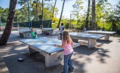 Jeunes personnes et adolescents en train de jouer au ping-pong sur une terrasse extérieure au camping Le Vieux Port, avec des tables de ping-pong en bois, entourés de végétation verdoyante et de structures de camping, ambiance conviviale et ensoleillée