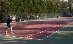 Groupe de trois hommes et une femme en tenue de sport, engagés dans un match de tennis sur un court en plein air au camping, avec des arbres et des tentes en arrière-plan, profitant de leurs vacances ensoleillées, montrant leur enthousiasme et leur concentration lors de la partie