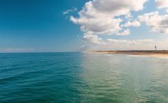 Vue depuis l’océan vers la plage de Messanges, avec sur la moitié gauche de l’image l’océan Atlantique s’étendant à perte de vue sous un ciel totalement dégagé sans nuages, et sur la moitié droite le phare de Messanges se détachant près du rivage sous un ciel partiellement couvert de nuages plus sombres au-dessus de la côte.