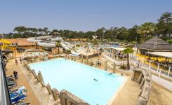 Vue panoramique en juin du parc aquatique XXL du camping 5 étoiles Le Vieux Port dans les Landes, avec plusieurs piscines extérieures chauffées, toboggans aquatiques géants, espace Aquasplash thématisé pirates avec crâne de pirate, pataugeoires pour enfants, jacuzzis, transats occupés par les vacanciers et grande pinède en arrière-plan sous un ciel bleu estival